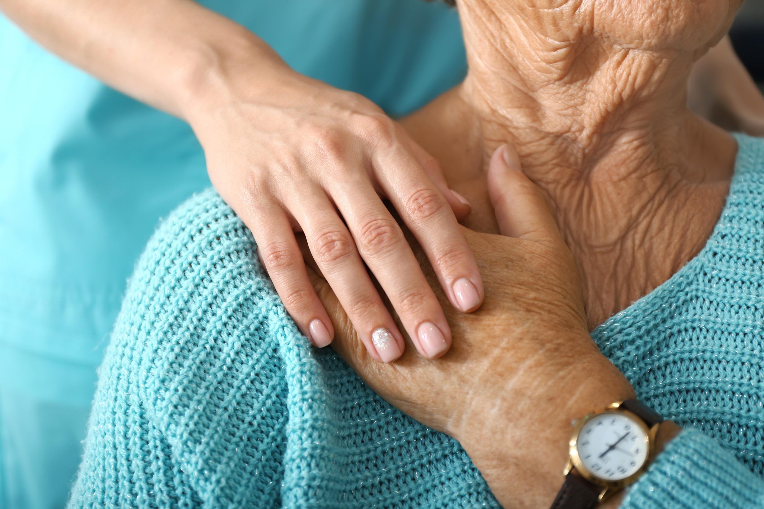 Elderly woman in teal coloured jumper with nurse touching her hand gently over the shoulder